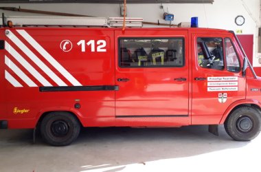 Ein rotes Feuerwehrauto mit der Notrufnummer ‚112‘ und weißen diagonalen Streifen an der Seite. Auf dem Fahrzeug sind Leitern und weitere Ausrüstung montiert. Es trägt die Aufschrift ‚Freiwillige Feuerwehr‘ und das Logo ‚Ziegler‘. Das Fahrzeug steht in einer Garage oder Feuerwache