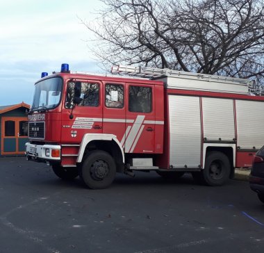 Ein großes, rotes Feuerwehrauto steht auf einem gepflasterten Platz. Auf der Vorderseite steht in weißen Großbuchstaben ‚FEUERWEHR‘. Im Hintergrund befinden sich Bäume und ein kleines Gebäude.