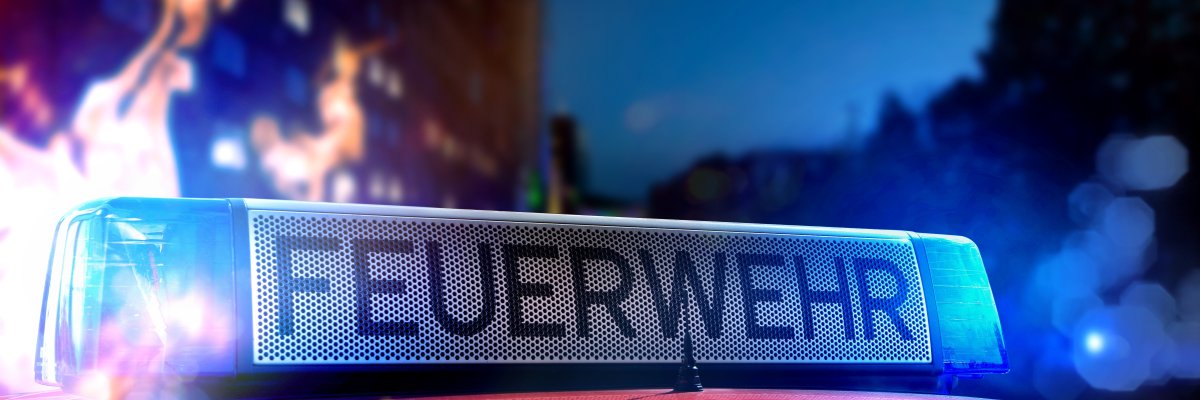 Oberseite eines Feuerwehrautos mit dem Schriftzug ‚FEUERWEHR‘ in großen Buchstaben. Im Hintergrund sind unscharfe Lichter und auf der linken Seite Flammen zu sehen – es wirkt wie ein nächtlicher Feuerwehreinsatz.