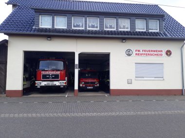 Feuerwehrhaus mit Ziegeldach. Im Erdgeschoss zwei Garagentore mit Feuerwehrfahrzeugen dahinter – links ein großes, rechts ein kleineres. Im Obergeschoss acht Fenster mit Gardinen. Rechts am Gebäude ein Schild mit der Aufschrift „FW. FEUERWEHR REIFFERSCHEID“ und zwei Logos.