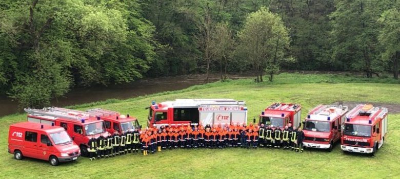 Eine Gruppe Feuerwehrleute steht in zwei Reihen vor sechs roten Feuerwehr- und Einsatzfahrzeugen. Die Szene spielt im Freien auf einer Wiese mit Wald im Hintergrund. Auf einem der mittleren Fahrzeuge steht ‚Freiwillige Feuerwehr‘ und ‚112
