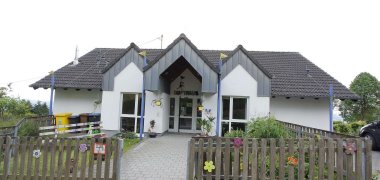 Kindergarten Luftikus Gebäudeansicht mit dem  Eingangsbereich umgeben mit einem Gartenzaun aus Holz dekoriert mit bunten Holzblüten 