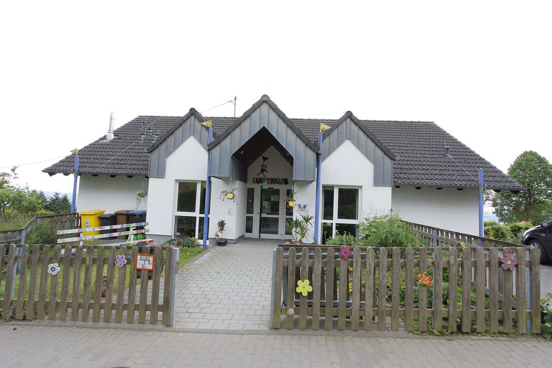 Kindergarten Luftikus Gebäudeansicht mit dem  Eingangsbereich umgeben mit einem Gartenzaun aus Holz dekoriert mit bunten Holzblüten 