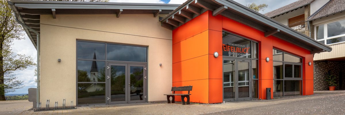 Modernes Gebäude mit beigen und orangefarbenen Fassaden. Große Glasfenster und Glastüren, davor eine Bank. Über dem Eingang steht „Eifelblick“. Im Hintergrund Bäume und ein teils bewölkter Himmel.