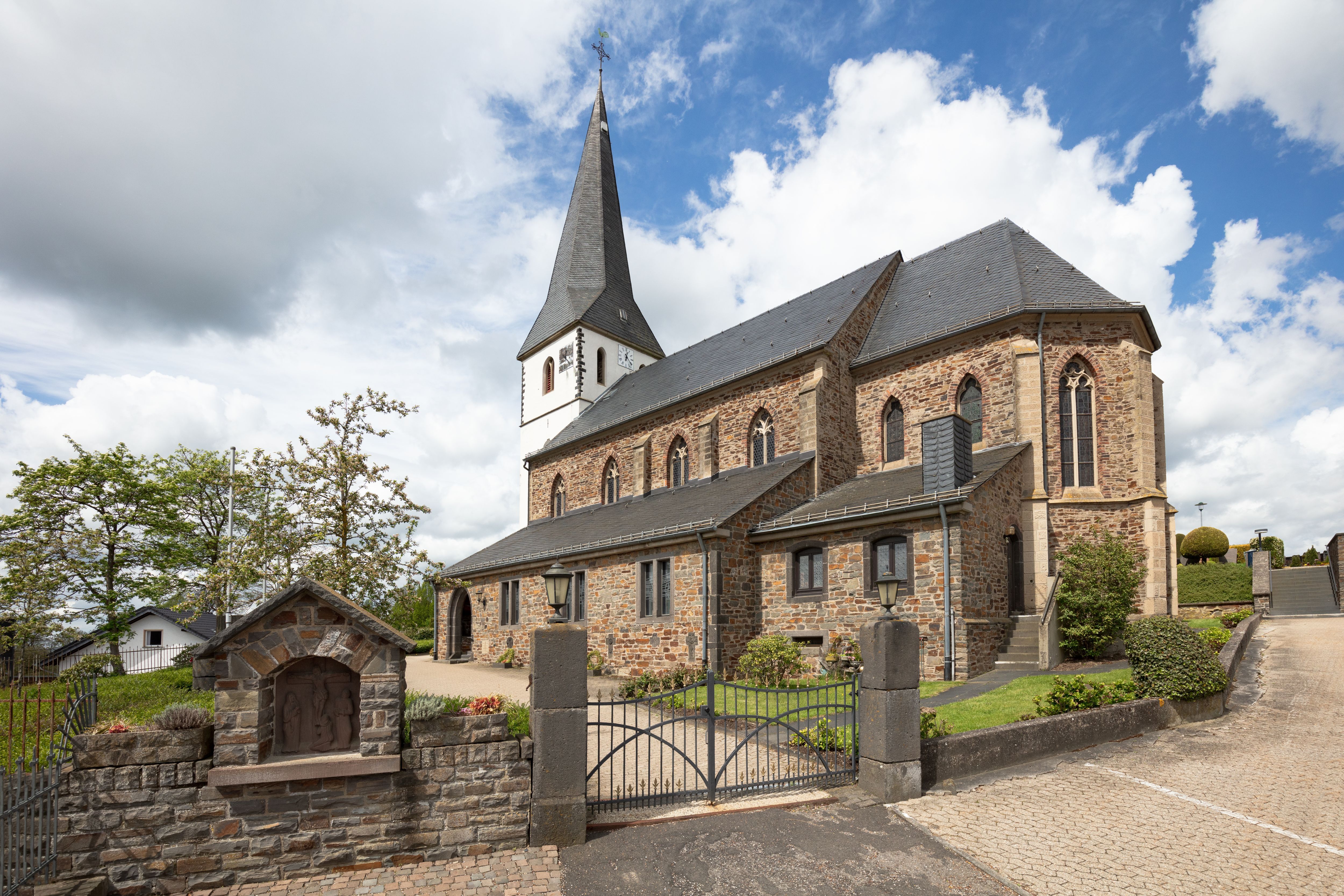 Kirche in Reifferscheid Steinkirche mit hohem, spitzem Turm und Uhr. Die Kirche hat Rundbogenfenster und ist von einer niedrigen Steinmauer mit Eisentor umgeben. Bäume und Büsche stehen rundherum, der Himmel ist teils bewölkt.