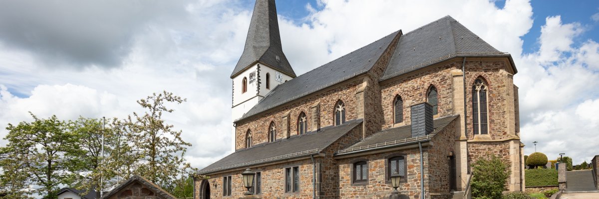 Steinkirche mit hohem, spitzem Turm und Uhr. Die Kirche hat Rundbogenfenster und ist von einer niedrigen Steinmauer mit Eisentor umgeben. Bäume und Büsche stehen rundherum, der Himmel ist teils bewölkt.