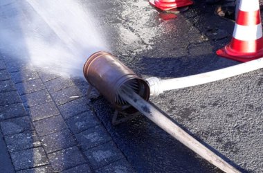 Ein Feuerwehr-Wasserschlauch ist an ein zylinderförmiges Gerät angeschlossen, das Wasser auf den Boden sprüht. Die Szene spielt sich auf einer gepflasterten Fläche ab. Im Hintergrund sind zwei Verkehrskegel und die Beine einer Person mit reflektierender Sicherheitskleidung zu sehen.