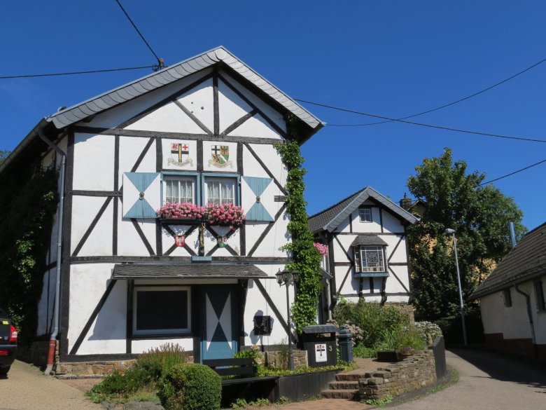 Zwei Fachwerkhäuser mit weißen Wänden und dunklen Holzbalken. Das vordere Haus hat blaue Fensterläden, Blumenkästen mit pinken Blumen und Efeu an der Wand. Über dem oberen Fenster sind zwei Wappen zu sehen. Im Hintergrund ein kleineres Fachwerkhaus. Die Szene spielt sich in einer schmalen Straße unter blauem Himmel ab.