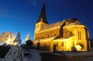 Kirche mit hohem Turm, nachts beleuchtet. Der Boden und Teile des Gebäudes sind mit Schnee bedeckt. Umgeben von verschneiten Bäumen und weiteren Gebäuden. Der Himmel ist dunkelblau.