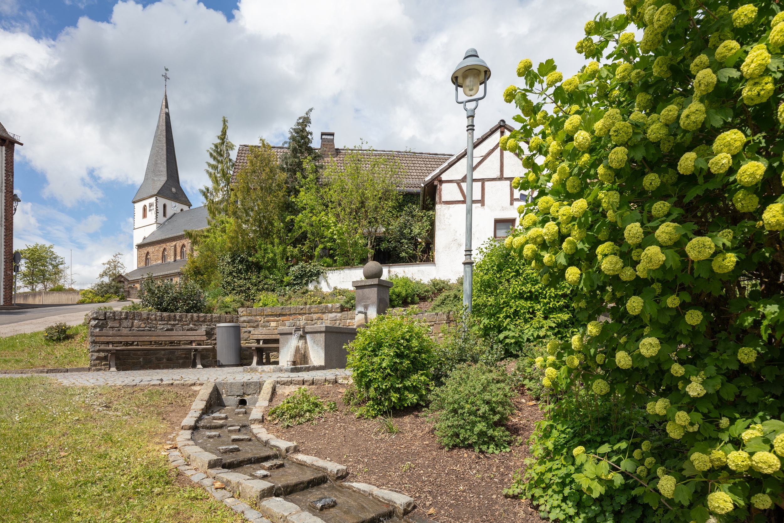 Dorfansicht mit Kirche und hohem Turm im Hintergrund. Im Vordergrund ein weißes Fachwerkhaus mit braunen Balken, umgeben von Bäumen und Sträuchern. Ein kleiner gemauerter Wasserlauf führt zu einem Brunnen mit Bänken und Mülleimern. Rechts blühende gelbgrüne Büsche, links eine Straßenlaterne. Der Himmel ist teils bewölkt mit blauen Abschnitten.