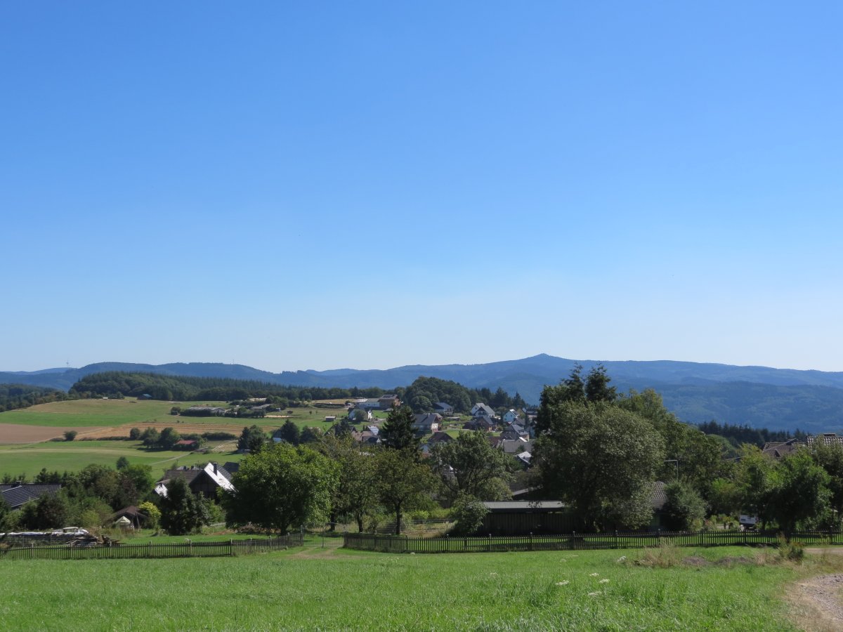 Ländliche Landschaft mit grünen Feldern im Vordergrund, einem kleinen Dorf mit Häusern und Bäumen in der Mitte und sanften Hügeln oder Bergen im Hintergrund. Darüber ein klarer blauer Himmel.